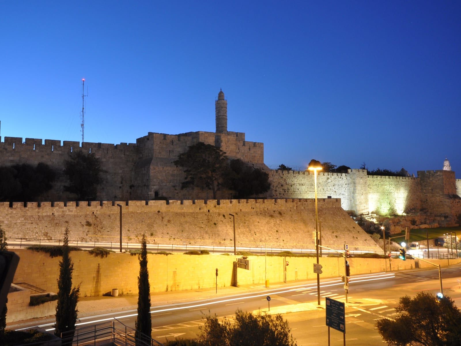 The Tower of David and the Old City walls of Jerusalem lit at dusk, above the road below.